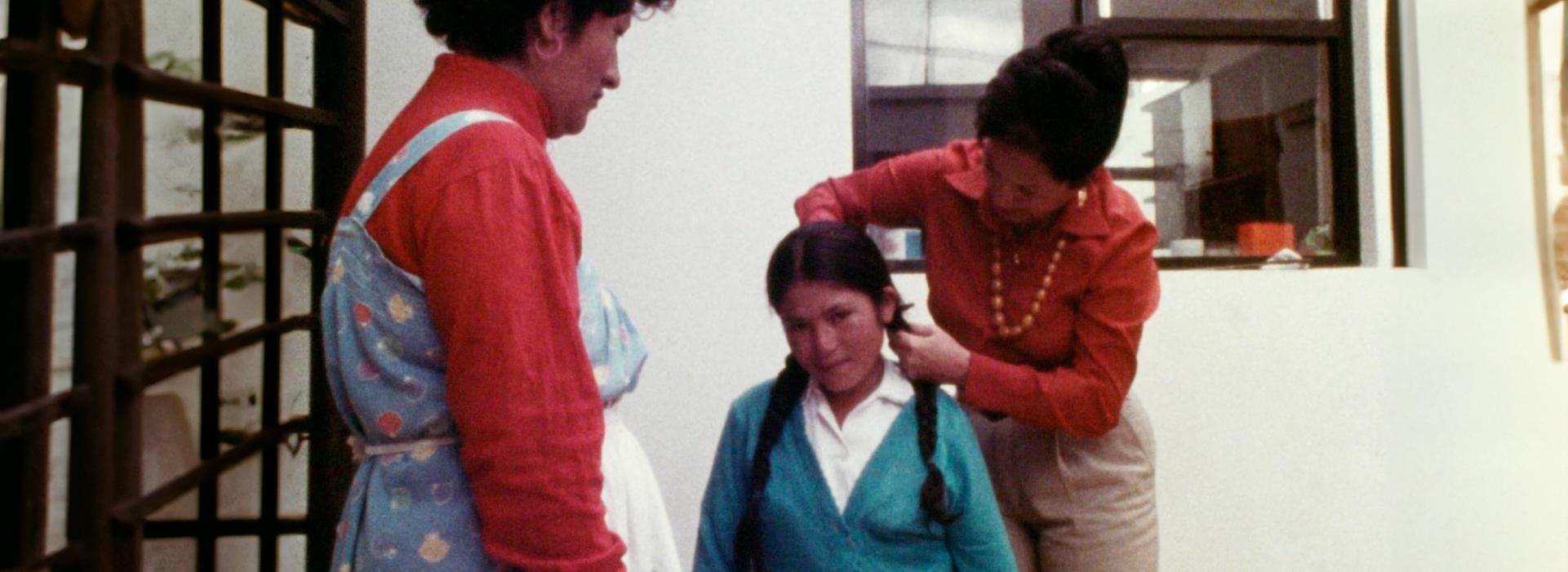 Hay dos mujeres y una niña. Una de las mujeres le corta las trenzas a la niña.