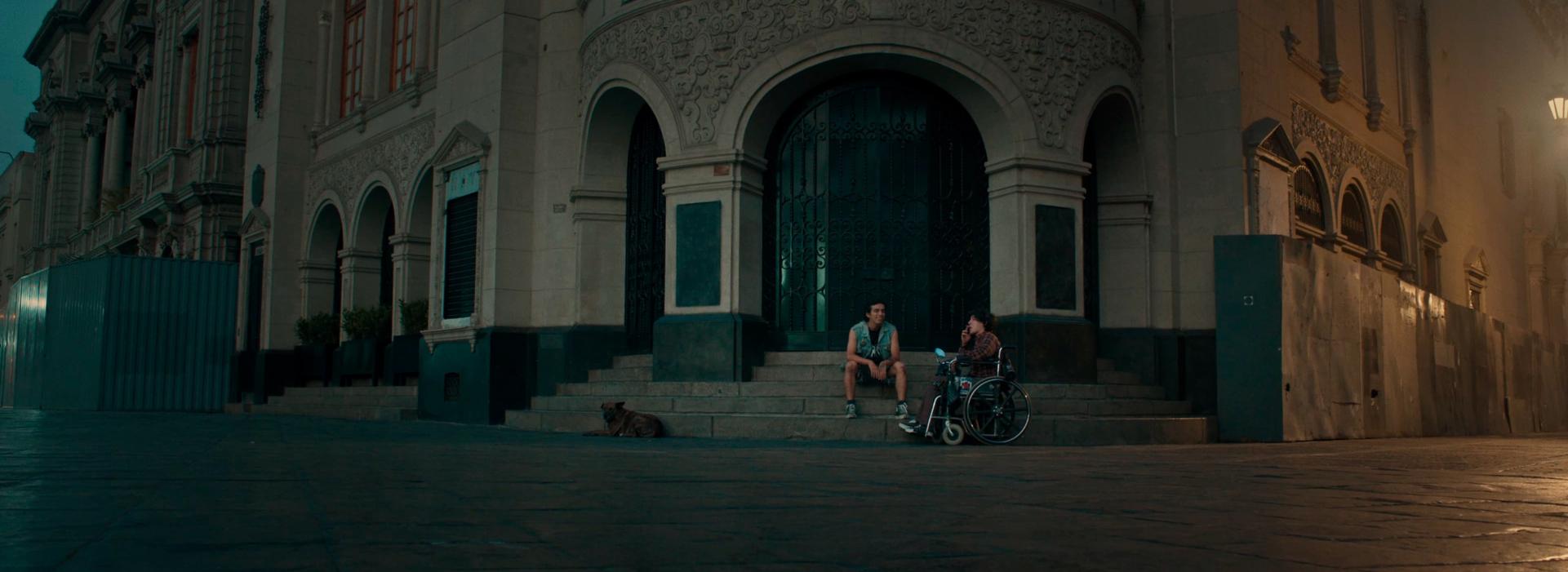 Un hombre está sentado en las escaleras del Teatro Colón, hablando con otro hombre en silla de ruedas. Hay un perro tumbado en las escaleras.