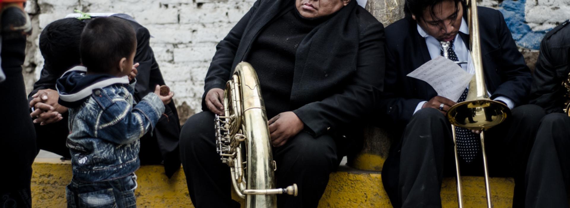 Tres hombres están sentados en la acera, uno tiene un cuerno, otro un trombón. Un niño los mira mientras come.