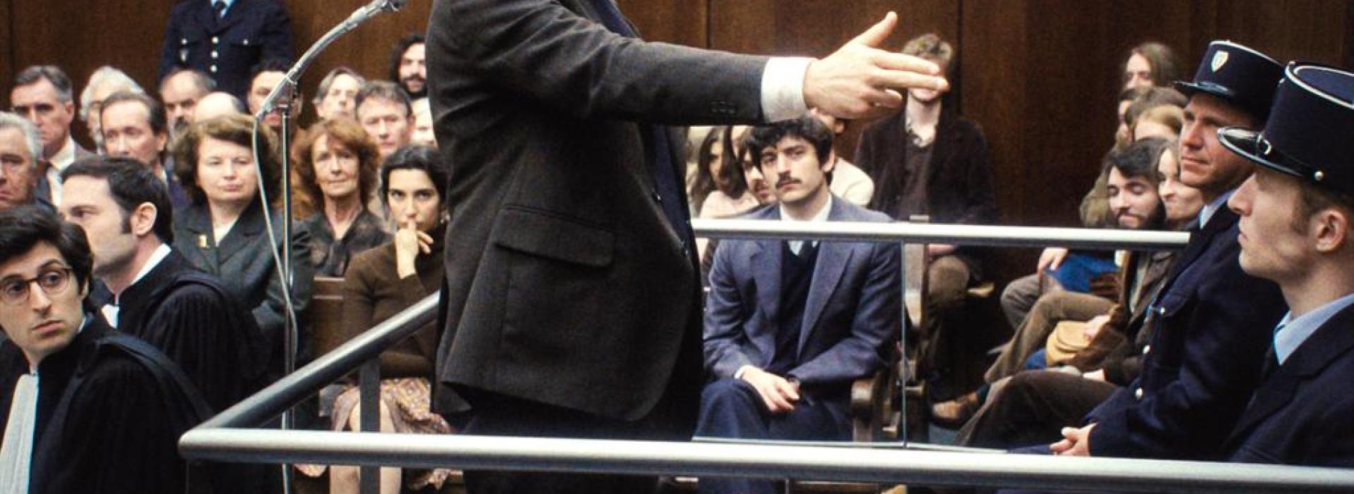 En una sala de juicios, un acusado en el estrado hace el gesto de una pistola con la mano hacia un policía. La sala está llena y observa la escena.